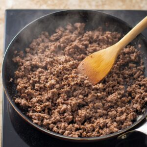 Easy Cheesy Taco Pasta Recipe 14 Ground beef being browned in a skillet with a wooden spoon stirring the meat. The beef is cooked until crumbly and browned, releasing its flavorful juices in preparation for the taco pasta dish.