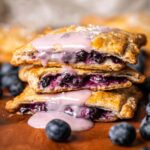 Blueberry Pop Tarts Close-up of baked blueberry hand pies stacked, golden flaky crust, juicy blueberry filling visible, topped with light pink glaze dripping, fresh blueberries around, shallow depth of field, warm natural light, high detail, professional food photography, 4k