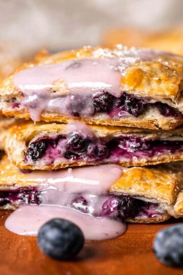 Healthy Blueberry Pop Tarts Recipe 3 Blueberry Pop Tarts Close-up of baked blueberry hand pies stacked, golden flaky crust, juicy blueberry filling visible, topped with light pink glaze dripping, fresh blueberries around, shallow depth of field, warm natural light, high detail, professional food photography, 4k