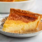 Buttermilk Pie A slice of buttermilk pie with a golden, flakey crust and smooth, custard-like filling. The pie is partially cut, revealing its creamy texture, and sits on the edge of a silver pie dish.