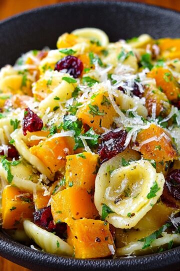Delicious Butternut Squash Pasta Salad Recipe 1 Butternut Squash Pasta Salad A black bowl filled with orecchiette pasta mixed with roasted butternut squash cubes, dried cranberries, chopped parsley, and topped with grated Parmesan cheese on a wooden surface.