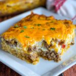 Cattle Drive Casserole A delicious serving of cattle drive casserole on a square white plate, showing the golden melted cheese on top and the hearty layers of beef, vegetables, and creamy sauce.