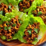 Chicken Lettuce Wraps Recipe Chicken lettuce wraps filled with a savory mixture of ground meat, cashews, and flavorful sauce, topped with fresh cilantro. The wraps are neatly arranged in vibrant green lettuce leaves on a wooden platter.