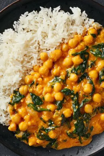 Delicious Chickpea Curry Recipe 1 Chickpea Curry A black ceramic plate with white basmati rice and creamy chickpea curry with spinach, served on a dark surface. The curry has a rich orange color and a glossy texture, with soft natural lighting enhancing the details.