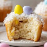 Coconut Easter Cupcakes A fluffy Easter cupcake with a coconut topping, sliced in half to reveal its soft, light interior, adorned with colorful Easter candy eggs on top