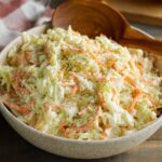 Coleslaw Recipe A bowl of creamy coleslaw made with shredded cabbage and carrots in a mayonnaise dressing, served with a wooden spoon on a rustic wooden table, with a hot dog visible in the background.