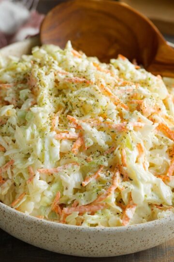 Healthy Coleslaw Recipe Recipe 1 Coleslaw Recipe A bowl of creamy coleslaw made with shredded cabbage and carrots in a mayonnaise dressing, served with a wooden spoon on a rustic wooden table, with a hot dog visible in the background.