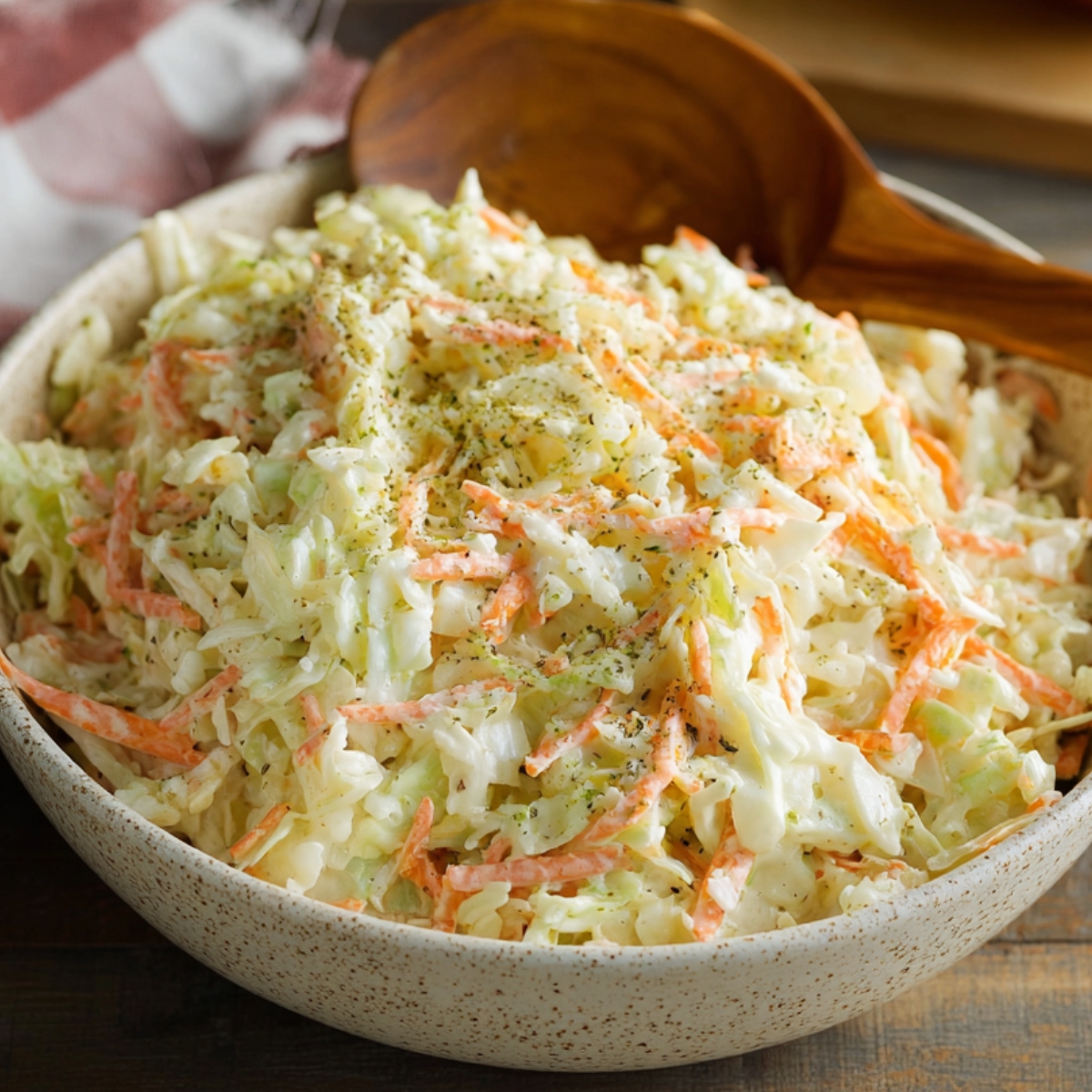 Coleslaw Recipe A bowl of creamy coleslaw made with shredded cabbage and carrots in a mayonnaise dressing, served with a wooden spoon on a rustic wooden table, with a hot dog visible in the background.