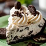 Cookies and Cream Pie A slice of Oreo cookies and cream pie on a green plate, topped with soft swirls of whipped cream, surrounded by whole Oreo cookies for an added touch