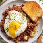 Corned Beef Hash A plate of homemade corned beef hash, topped with a sunny-side-up egg and served with toasted bread.