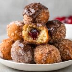 Donut Holes A plate of various homemade donut holes stacked together, some coated with cinnamon sugar, others with chocolate ganache or jam filling, with a bite taken out of one.