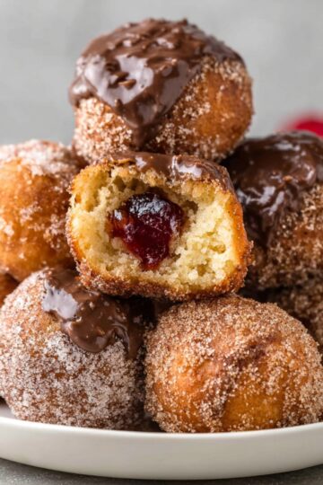 Best Homemade Donut Holes Recipe 1 Donut Holes A plate of various homemade donut holes stacked together, some coated with cinnamon sugar, others with chocolate ganache or jam filling, with a bite taken out of one.