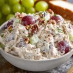 Fruity Chicken Salad Creamy chicken salad with red grapes, chopped celery, and pecans in a white bowl, topped with black pepper, served on a light surface with fresh grapes and sandwich bread in the background.