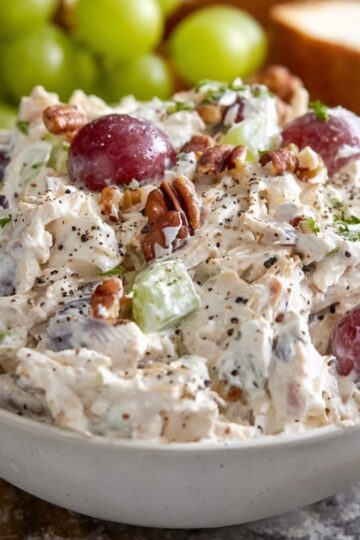 The Best Fruity Chicken Salad Recipe 2 Fruity Chicken Salad Creamy chicken salad with red grapes, chopped celery, and pecans in a white bowl, topped with black pepper, served on a light surface with fresh grapes and sandwich bread in the background.