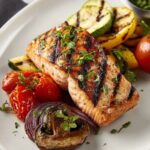 A beautifully plated grilled salmon fillet served with grilled vegetables like zucchini, tomatoes, and mushrooms, garnished with fresh herbs