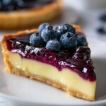 Lemon Blueberry Tart A beautifully sliced blueberry tart showing its layers. The tart has a golden, flaky crust with a luscious blueberry filling on top. A slice has been removed, revealing the smooth, fruit-filled interior.