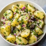 Mediterranean Potato Salad A bowl of Mediterranean-style potato salad with tender, boiled potatoes, garnished with fresh parsley, red onion, and black olives. The salad is lightly seasoned with pepper, creating a savory and refreshing dish perfect for a summer meal.