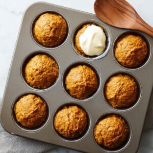 A person is spooning pumpkin muffin batter into muffin cups in a metal muffin tin, preparing for baking.