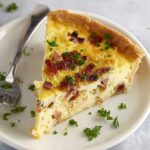 Quiche Lorraine A slice of Quiche Lorraine served on a white plate with bacon and cheese filling, garnished with parsley.