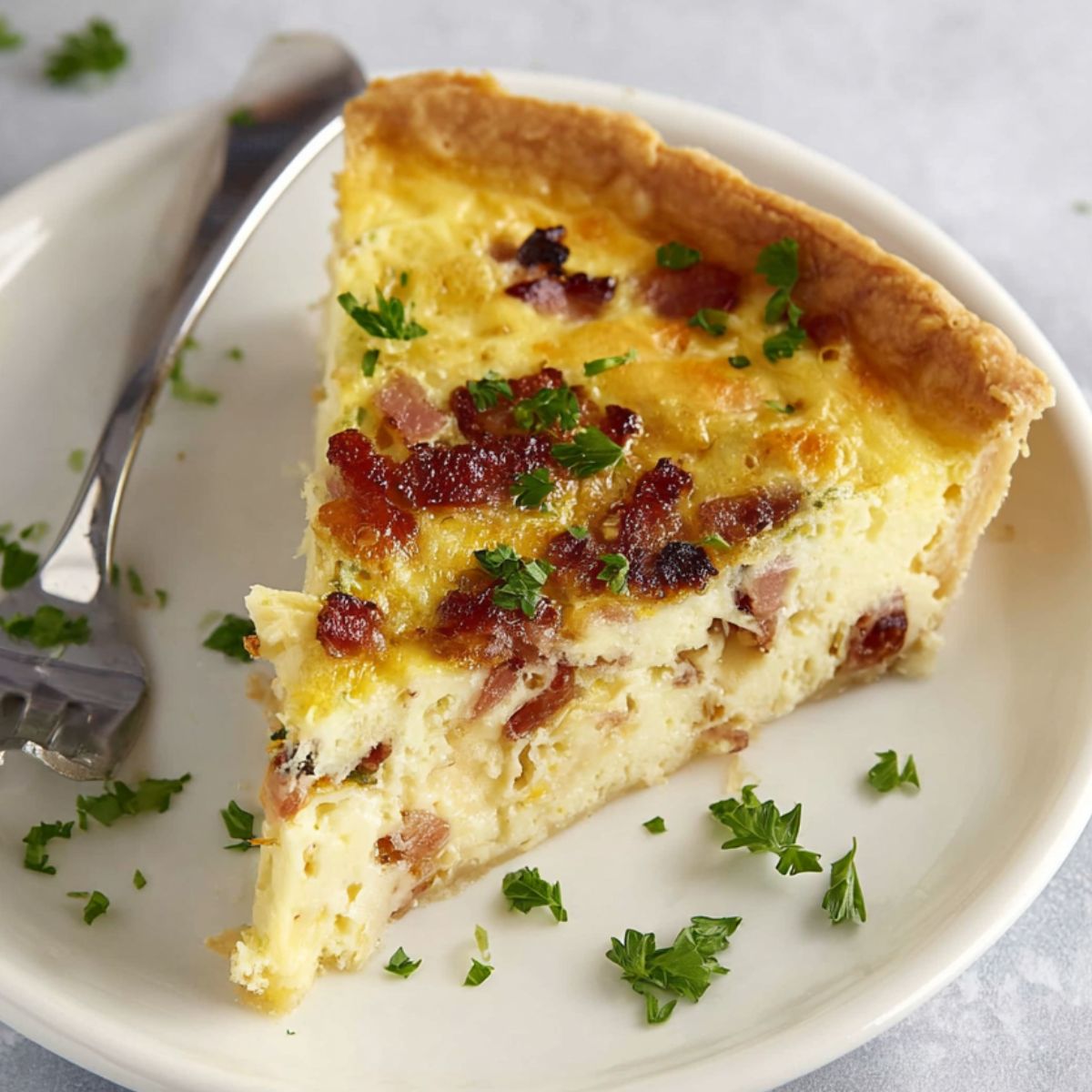 Quiche Lorraine A slice of Quiche Lorraine served on a white plate with bacon and cheese filling, garnished with parsley.