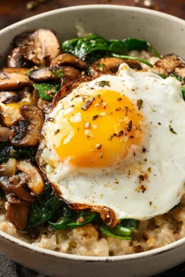 Savory Oatmeal A rustic food photography shot of a savory oatmeal bowl topped with a sunny-side-up egg, sautéed mushrooms, and wilted spinach, served in a white ceramic bowl on a dark wooden table, natural light, shallow depth of field, high detail, warm tones, professional food styling.