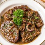 Smothered Cube Steak A perfectly plated dish of smothered cube steak, garnished with fresh parsley, with a side of bread and scattered herbs
