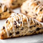 Sourdough Chocolate Chip Scones Close-up of freshly baked chocolate chip scones drizzled with vanilla glaze, golden brown and flaky texture, soft interior with visible chocolate chunks, on a white surface, natural soft lighting, shallow depth of field, food photography, high resolution, cozy bakery style