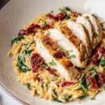Tuscan Chicken Orzo A close-up of a plate with creamy Tuscan chicken orzo, featuring a sliced chicken breast on top. The dish includes vibrant spinach, sun-dried tomatoes, and a rich, creamy sauce. A fork and herbs add to the elegant presentation