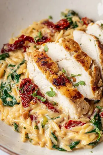 Tuscan Chicken Orzo A close-up of a plate with creamy Tuscan chicken orzo, featuring a sliced chicken breast on top. The dish includes vibrant spinach, sun-dried tomatoes, and a rich, creamy sauce. A fork and herbs add to the elegant presentation