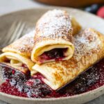 blintzes Recipe A delicious serving of blintzes, filled with a sweet or savory mixture, dusted with powdered sugar, and resting on a plate with berry sauce underneath.