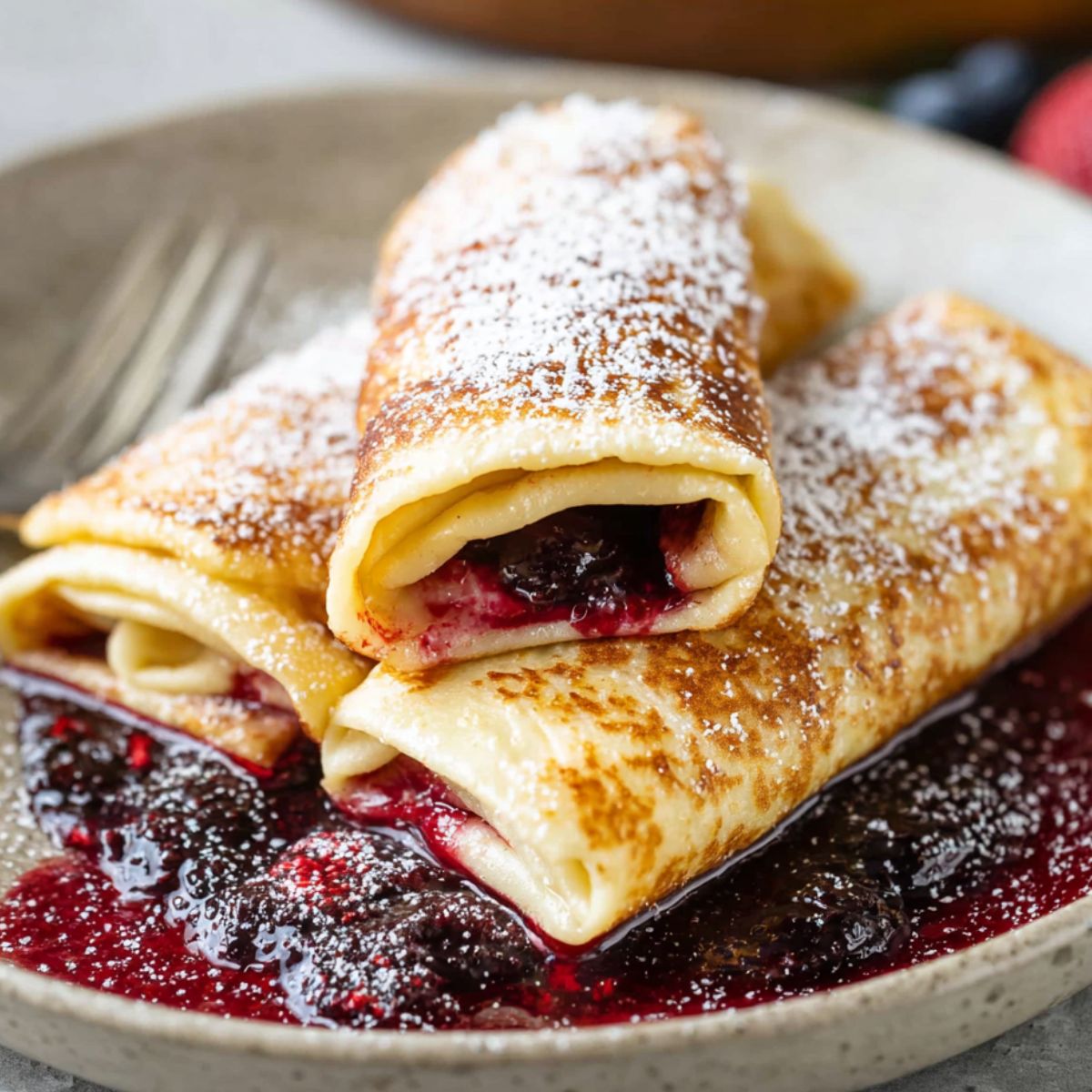 blintzes Recipe A delicious serving of blintzes, filled with a sweet or savory mixture, dusted with powdered sugar, and resting on a plate with berry sauce underneath.