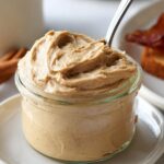 cinnamon butter Close-up of a small glass jar filled with whipped cinnamon honey butter, a spoon resting inside, with blurred bacon and cinnamon sticks in the background.