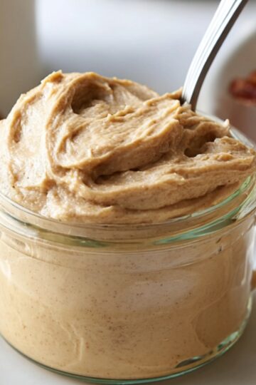 Delicious Cinnamon Butter Recipe 1 cinnamon butter Close-up of a small glass jar filled with whipped cinnamon honey butter, a spoon resting inside, with blurred bacon and cinnamon sticks in the background.