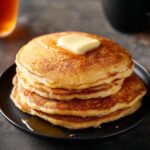 sweet potato pancakes A stack of golden-brown pancakes served on a black plate, with melted butter and syrup on the side. The pancakes have a fluffy and light texture, showcasing a perfect breakfast setup.