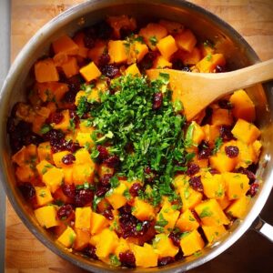 Delicious Butternut Squash Pasta Salad Recipe 15 A pot filled with diced butternut squash, dried cranberries, and chopped parsley being stirred with a wooden spoon on a stovetop.