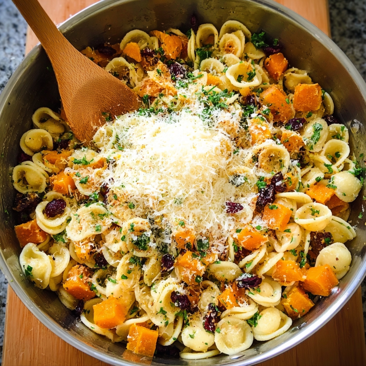 Delicious Butternut Squash Pasta Salad Recipe 12 A large metal pot of orecchiette pasta combined with roasted butternut squash, dried cranberries, and parsley, topped with a pile of freshly grated Parmesan cheese and a wooden spoon inside.