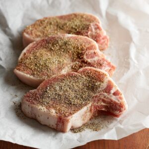 Easy Smothered Pork Chops Recipe 15 Raw pork chops coated with seasoning, resting on parchment paper, awaiting the next step in the cooking process.