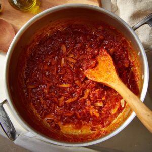 A wooden spoon stirring caramelized tomato paste in a pot, creating a deep red, rich color as the paste blends with the cooking ingredients.