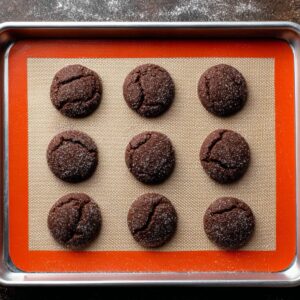 Delicious Andes Mint Chocolate Cookies Recipe 17 Nine round chocolate sugar cookies arranged in a 3×3 grid on a metal baking sheet lined with a beige silicone mat with an orange border, set on a dark, textured surface.