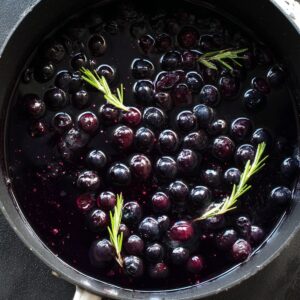 Healthy Blueberry Pop Tarts Recipe 14 Top-down food photography of a black saucepan filled with dark blueberry compote, whole blueberries floating in deep purple syrup, small sprigs of fresh rosemary, glossy texture, natural soft daylight, high detail, moody tones, minimal background, realistic, 4k, professional food styling