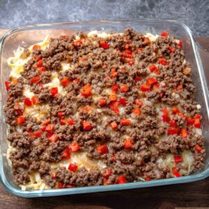 A casserole dish with a cooked beef mixture, featuring ground beef, diced red peppers, and onions, layered on top of a base, ready to be baked with cheese and other ingredients.