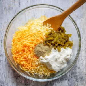 A clear glass mixing bowl containing shredded cheddar cheese, sour cream, green chilies, and seasoning powder, ready to be mixed into a delicious casserole for the cattle drive dish.