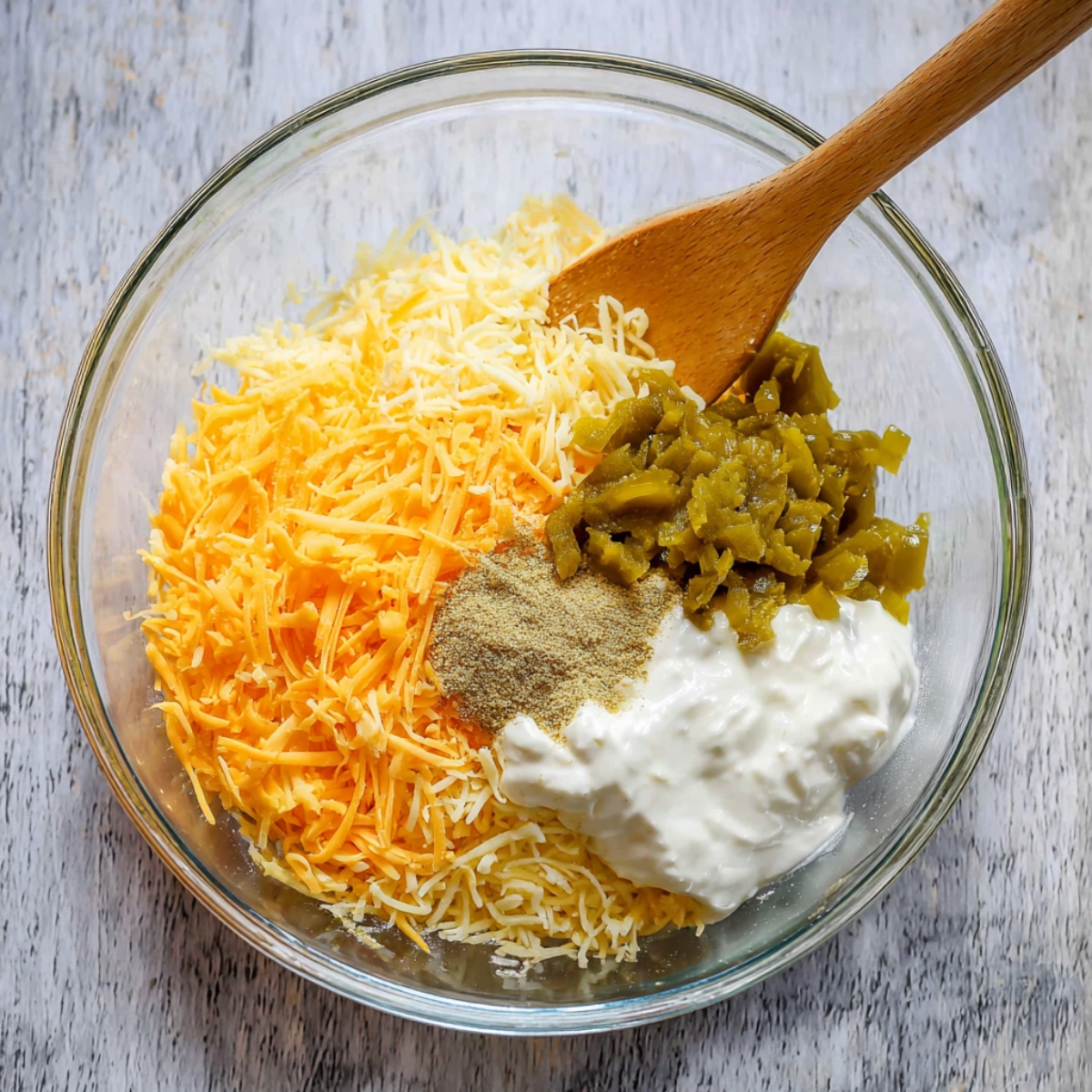 Delicious Cattle Drive Casserole Recipe 12 A clear glass mixing bowl containing shredded cheddar cheese, sour cream, green chilies, and seasoning powder, ready to be mixed into a delicious casserole for the cattle drive dish.
