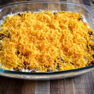 A casserole dish filled with a hearty cattle drive casserole topped with a generous layer of shredded cheddar cheese, ready to be baked. The casserole looks creamy with savory layers of ingredients.