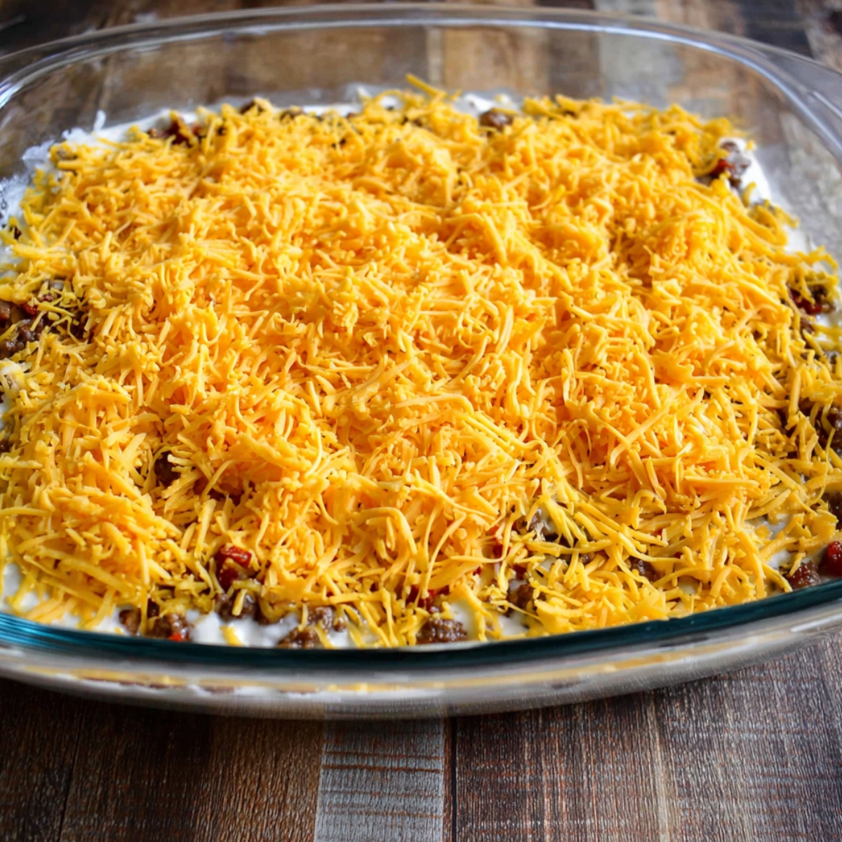 Delicious Cattle Drive Casserole Recipe 13 A casserole dish filled with a hearty cattle drive casserole topped with a generous layer of shredded cheddar cheese, ready to be baked. The casserole looks creamy with savory layers of ingredients.