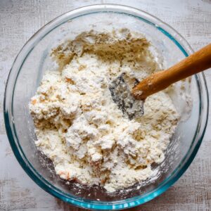 A glass bowl with a thick mixture of ingredients for the casserole, including flour and other dough elements, mixed together with a spatula, preparing for assembly in a casserole.