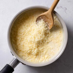 Best Cauliflower Gratin Recipe 14 A saucepan containing a creamy mixture with grated cheese being stirred in. The cheese is melting and combining into the sauce, creating a rich and cheesy texture