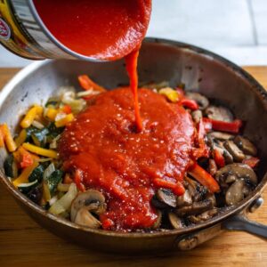 "Crushed tomatoes being poured into a pan with sautéed vegetables, forming the base for a rich and savory chicken cacciatore sauce.