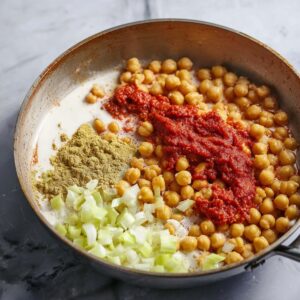 Delicious Chickpea Curry Recipe 16 A close-up of chickpeas, tomato paste, and coconut milk being added to a pan with chopped leeks, forming the base for a flavorful curry or stew.