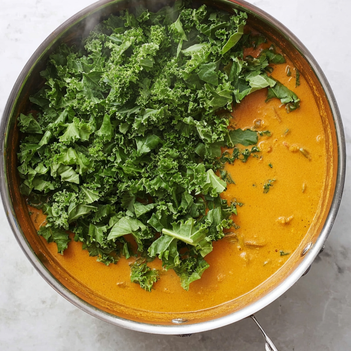 Delicious Chickpea Curry Recipe 12 A top-down view of a stainless steel pan with a curry sauce on one side and freshly added chopped kale on the other. The vibrant green kale contrasts with the orange sauce, and steam rises from the pan, set against a white marble surface.